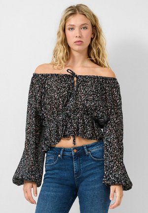 Jonge vrouw met golvend blond haar, gekleed in een zwart bloemenblouse met blote schouders en een blauwe spijkerbroek, staand tegen een effen achtergrond.