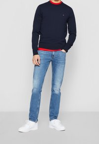 Marineblaues Sweatshirt mit rotem Kragen, über einem roten Hemd getragen, kombiniert mit hellblauen Jeans und weißen Turnschuhen. Einfaches Design, lässiger Look.
