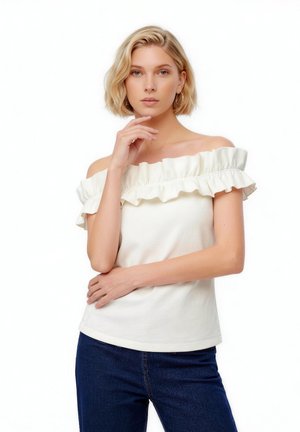 Plavuša s kratkom kosom, odjevena u bijelu bluzu s naborima bez ramena i tamnoplave traperice, pozira s rukom blizu brade na bijeloj pozadini.