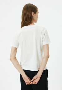 Hvid bomulds t-shirt, korte ærmer, rund halsudskæring og en afslappet pasform. Modellen står med hænderne foldet bag ryggen.