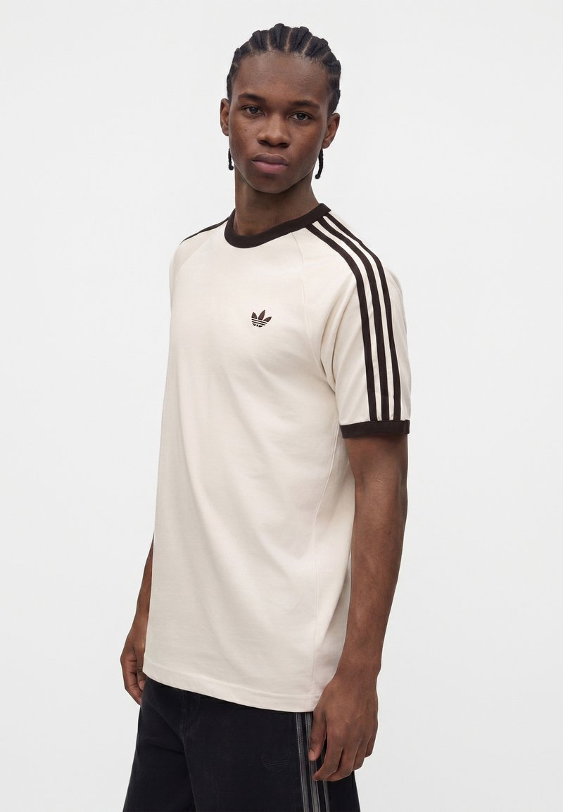 Junger Mann mit geflochtenem Haar, der ein cremefarbenes Adidas T-Shirt mit schwarzen Streifen an den Ärmeln und Logo trägt, steht vor einem schlichten weißen Hintergrund.