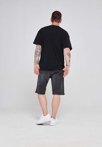 Juodas trumpomis rankovėmis marškinėliai, laisvas kirpimas, derinami su tamsiai pilkais džinsiniais šortais. Modelis vilki baltus sportinius batus, demonstruodamas kasdienį įvaizdį.