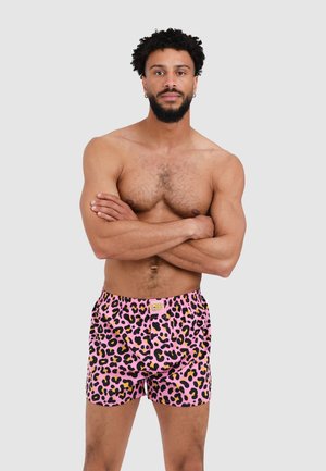 Shirtless man with curly hair and beard standing with arms crossed, wearing pink leopard print boxer shorts against a plain background.
