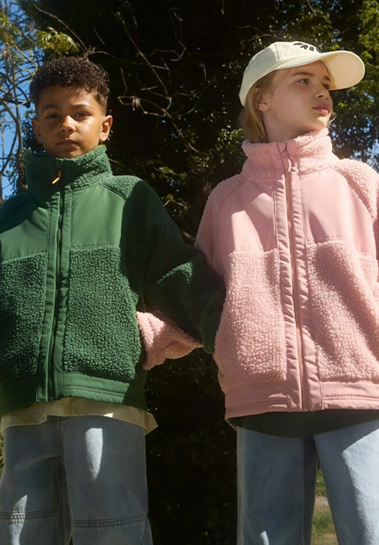 To børn holder hinanden i hånden, iført fuldforede jakker. Den ene jakke er grøn fleece med en glat overflade, mens den anden er lyserød fleece med teksturerede detaljer.