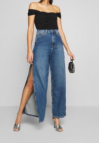 Korkeavyötäröiset, leveälahkeiset siniset farkut, joissa on sivuaukot, yhdistetty mustaan olkaimettomaan toppiin ja hopeisiin korkokenkiin, luoden rennon ilmeen.