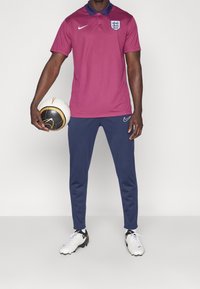 Bordowa koszulka polo z granatowym kołnierzykiem z logo Anglii, w połączeniu z granatowymi spodniami i czarno-białymi butami piłkarskimi, trzymająca czarno-białą piłkę nożną.