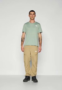 Világos zöld, rövid ujjú póló, bézs cargo nadrág, fekete cipő. Kiemelkedő jellemzői közé tartozik a The North Face logó és a cipzáros zsebek.