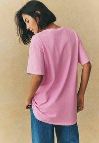 Rozā kokvilnas T-krekls ar īsām piedurknēm un brīvu piegriezumu, kopā ar ziliem džinsu biksēm. Audums izskatās mīksts ar gludu tekstūru.