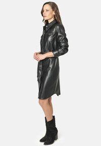 Vestido camisero negro de piel sintética con mangas largas, cierre de botones en la parte delantera, bolsillos y cintura ceñida. Combinado con botas de cowboy negras con adornos.