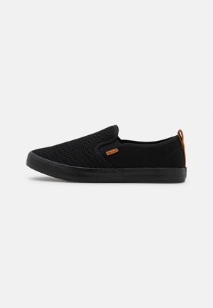 Svarta slip-on skor med mesh-överdel och solida svarta detaljer. Har en brun etikett och en gummisula. Texturerad yta.