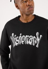 Svart hettegenser med en stor, teksturert hvit "Visionary" grafikktrykk på forsiden. Stoffet ser mykt ut med en klassisk rund hals.