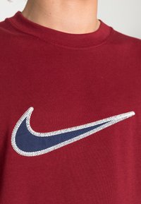 Maroon cotton T-shirt featuring a navy blue Nike swoosh logo with white stitching, round neckline, and short sleeves.
