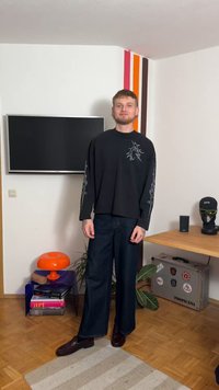 Melna garām piedurknēm krekls ar grafisku dizainu, kombinēts ar plaša piegriezuma tumšām džinsa biksēm un brūnām kurpēm. Aiz muguras redzama krāsaina lampa un sienas dekors.