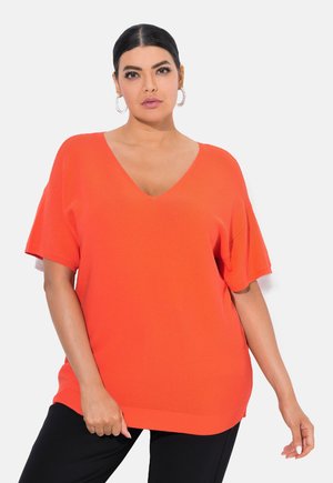 Felpa oversize arancione brillante con scollatura a V e maniche corte, realizzata in tessuto a costine; abbinata a pantaloni neri.