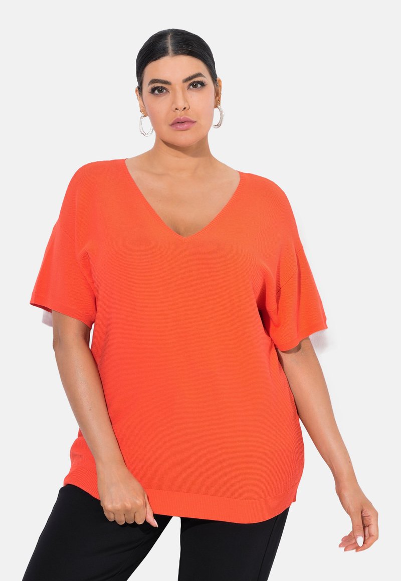 Světlý oranžový oversized top s V výstřihem a krátkými rukávy, vyrobený z jemné texturované látky; v kombinaci s černými kalhotami.