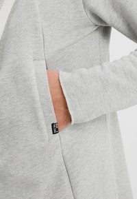 Main d'une personne dans la poche latérale d'un hoodie gris clair avec une petite étiquette noire indiquant "WEAR THE CODE."