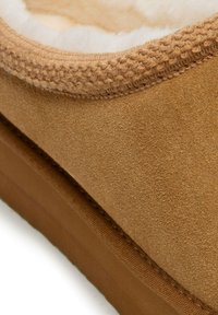 Zapatilla de terciopelo marrón con un forro interior suave y peludo. Presenta un borde texturizado y cosido, y una suela plana de goma para mayor agarre.