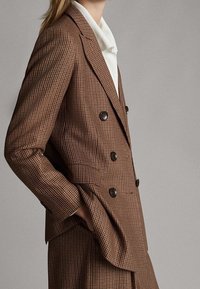 Brązowa w kratę marynarka dwurzędowa z dużymi czarnymi guzikami, dopasowany krój i klapy w szpic. Gładki materiał o wyraźnej sylwetce.