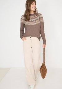 Maglione marrone con un motivo geometrico crema, pantaloni beige a gamba larga e una borsa in suede marrone. Sneakers bianche completano l'outfit.