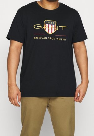 Mand iført sort T-shirt med gul og rød GANT-logo og teksten "American Sportswear", kombineret med khaki bukser, stående mod hvid baggrund.