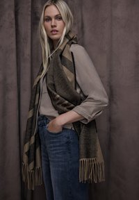 Écharpe rayée marron et beige avec bords effilochés drapée sur une chemise marron clair, associée à un jean bleu. Textures douces et couleurs complémentaires.