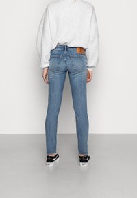Persona che indossa jeans slim-fit azzurri chiari, un sweatshirt bianco e scarpe ginniche bianco e nere, in piedi di fronte a un muro bianco uniforme.