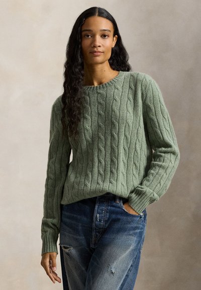 Donna con lunghi capelli ricci e scuri che indossa un maglione verde a trecce e jeans blu consumati, in piedi con una mano in tasca.