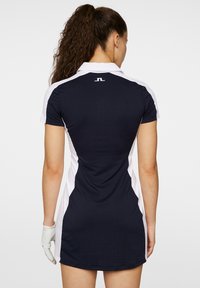 Marinblå och vit klänning med kort ärm, figurnära passform, krage och logotyp på baksidan. Slät textur och knälång.