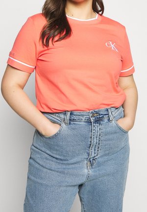 Nainen, jolla on päällään koralli lyhythihainen t-paita valkoisella reunuksella ja vaaleansiniset korkeavyötäröiset farkut, kädet taskuissa, näkyvissä vain ylävartalo.