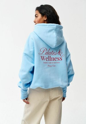 Kobieta ubrana w jasnoniebieską bluzę z kapturem, z napisem '"Pilates & Wellness"' i powiązanym tekstem wydrukowanymi na czerwono z tyłu, stoi na tle jednolitego tła.
