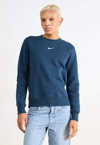 Mørkeblå hettegenser med rund hals, ribbede mansjetter og nederkant; har en liten hvit Nike-logo på brystet; laget av bomullsblandingsmateriale.