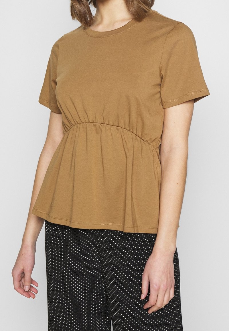 Femme portant un haut marron à manches courtes avec un détail froncé à la taille et un pantalon noir à petits pois blancs.