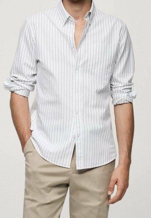 Mann trägt ein weißes langärmeliges Hemd mit dünnen vertikalen Streifen und beiger Hose, Hemdärmel hochgekrempelt, eine Hand in der Tasche.