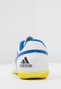 Vit Adidas sportssko med blå accenter och en gul sula, sedd bakifrån, som visar varumärkesloggen.