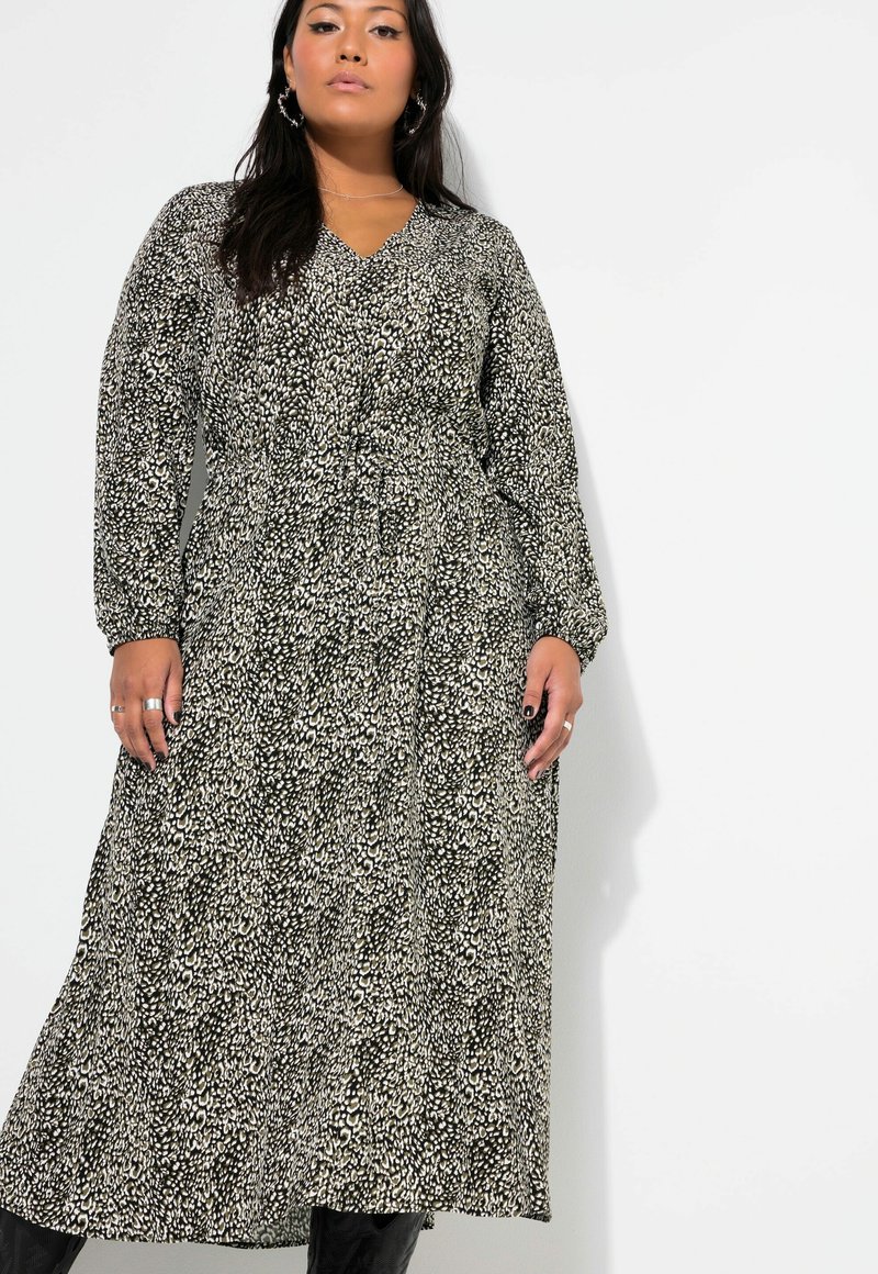 Schwarz-weiß gemustertes Maxikleid mit langen Ärmeln, V-Ausschnitt, tailliertem Schnitt und fließendem Rock. Getragen mit schwarzen Stiefeln und Accessoires.