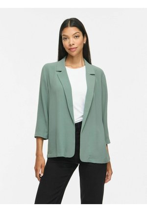 Kvinde med langt mørkt hår iført en salviegrøn åben blazer, hvid top og sorte bukser, stående foran en ensfarvet hvid baggrund.