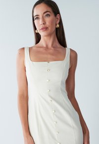 Robe crème sans manches avec encolure carrée et boutons sur le devant, confectionnée dans un tissu lisse.