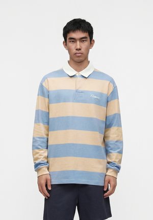 Jeune homme portant un maillot de rugby à manches longues avec de larges rayures horizontales bleues et beiges et un short foncé, debout devant un fond uni.
