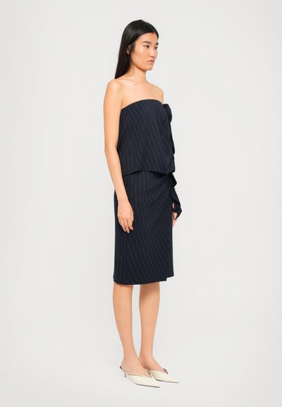 Vrouw in een marineblauwe strapless pinstripe jurk met ruches, die witte puntige lage hakken draagt tegen een effen achtergrond.