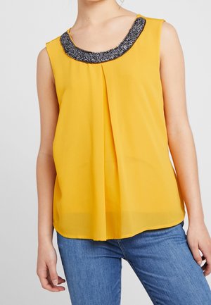 Femme portant un chemisier jaune sans manches avec un détail de col perlé noir et blanc et un jean bleu, debout devant un fond uni.