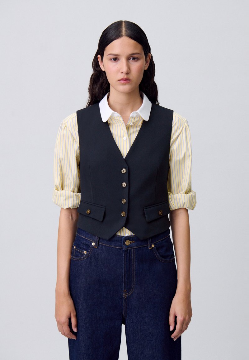 Una giovane donna con lunghi capelli scuri indossa un gilet nero su una maglietta a righe gialle e jeans scuri, in piedi contro uno sfondo semplice.