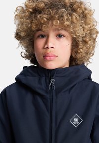 Granatowa kurtka z kapturem z zamkiem błyskawicznym, z logo naszytym na przodzie oraz sznurkiem do regulacji. Materiał wygląda na wodoodporny i ma gładką teksturę.