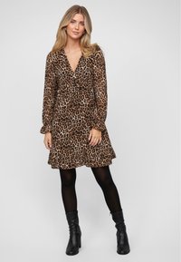 Kleid im Leopardenmuster mit langen Ärmeln, tiefem V-Ausschnitt und Rüschensaum. Hergestellt aus leichtem Stoff, kombiniert mit schwarzen Stiefeletten.