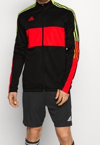 Chaqueta con cremallera negra y roja de mangas largas, con detalles en verde y tres franjas. Combinada con pantalones cortos grises y calcetines en blanco y negro.