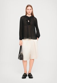 Svart, gjennomsiktig bluse med volangdetaljer, kremfargede, pliserte shorts, svarte loafers og en mørk håndveske. Enkelt og lett stoff.