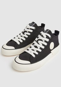 Černé plátěné tenisky s bílými tkaničkami, kulatou nášivkou loga a kontrastní gumovou špičkou. Texturovaná bílá gumová podrážka s vroubkovaným vzorem.