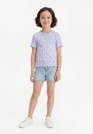 Ung pige står iført en lilla kortærmet skjorte med hvide tusindfryd, lyse denimshorts og hvide sneakers mod en enkel baggrund.