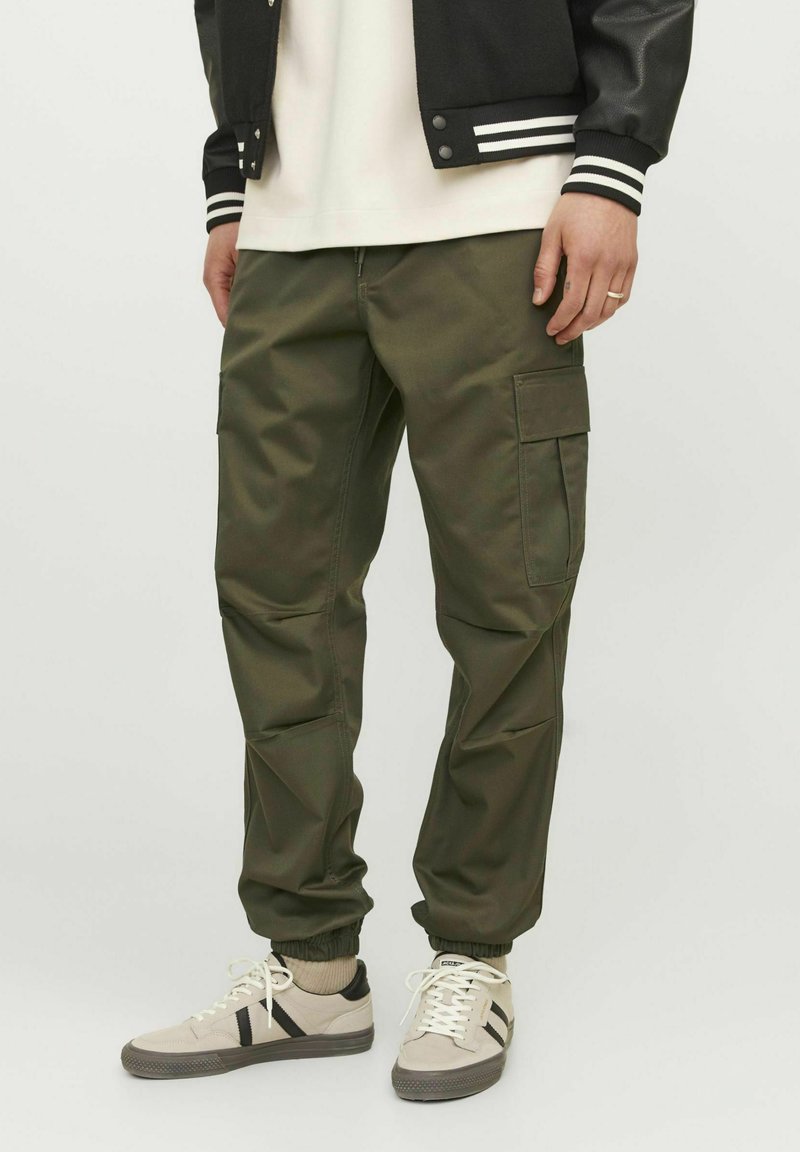 Pantalones cargo verde oliva con piernas ajustadas, puños elásticos y bolsillos laterales, combinados con zapatillas de color claro y una chaqueta de universidad negra.