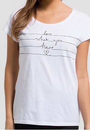 Vrouw draagt een wit T-shirt met zwarte handgeschreven tekst "love what you have" en een klein hartje onder de tekst.