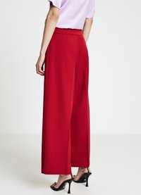 Pantalon rouge à jambes larges en tissu lisse, avec une taille haute, des plis et une coupe ajustée. Porté avec des sandales noires à talons hauts.